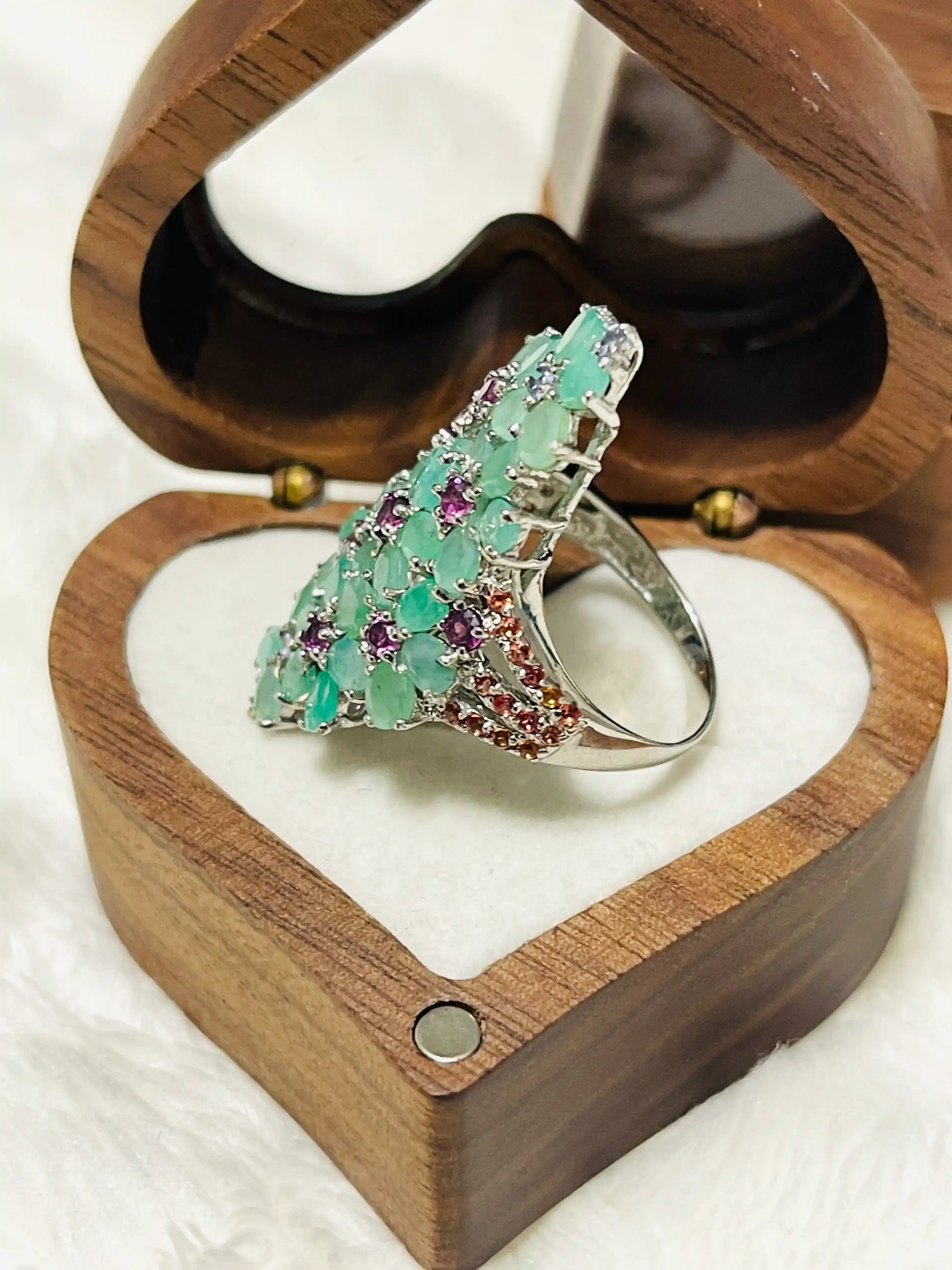 Profile of a striking cocktail ring featuring numerous emeralds and purple amethyst stones, elegantly displayed in a box.