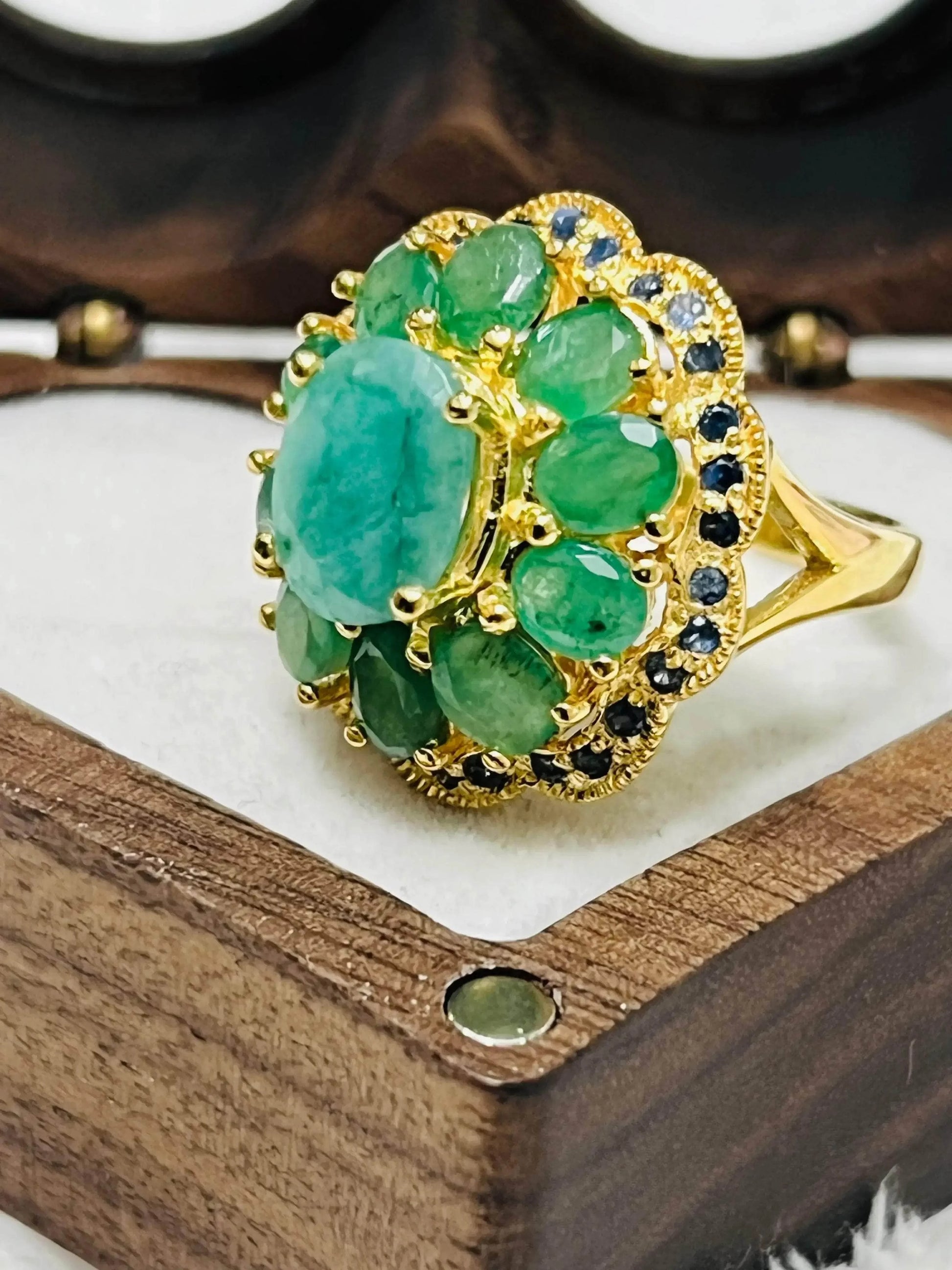Gold cocktail ring with oval emerald center, surrounded by green emeralds and blue sapphires in a vintage-style setting.