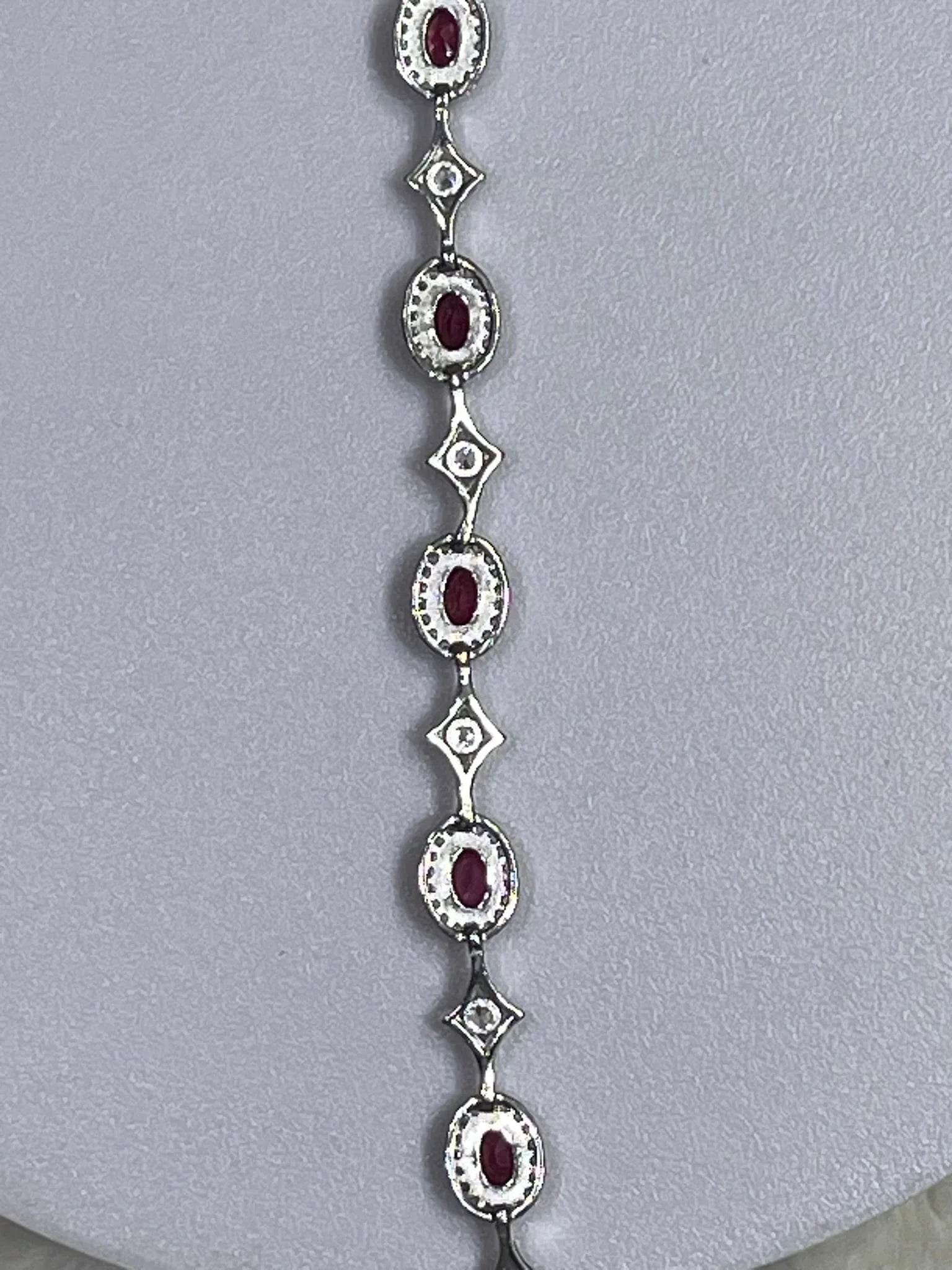 Vertical shot of a silver bracelet featuring a series of oval rubies and diamond-studded links, on a neutral background.