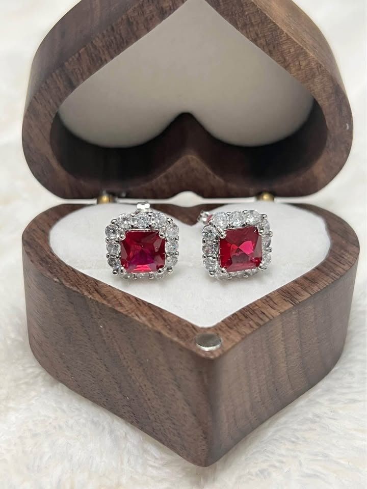 Front view of two sterling silver ruby stud earrings with halo setting, nestled in a wooden heart box.