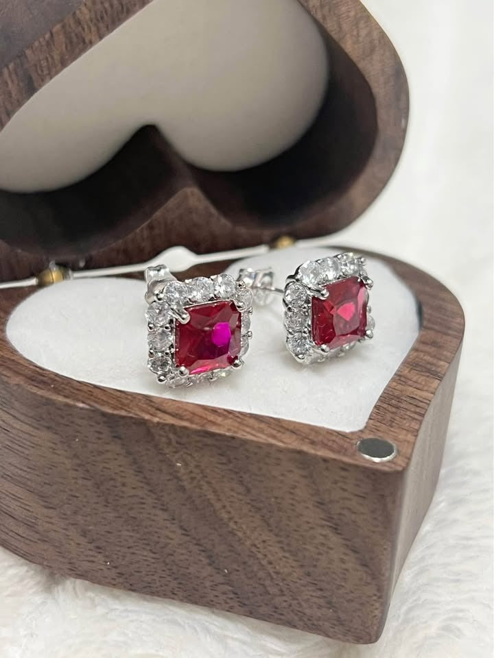Pair of sterling silver ruby stud earrings with halo in a heart-shaped wooden box, side view.