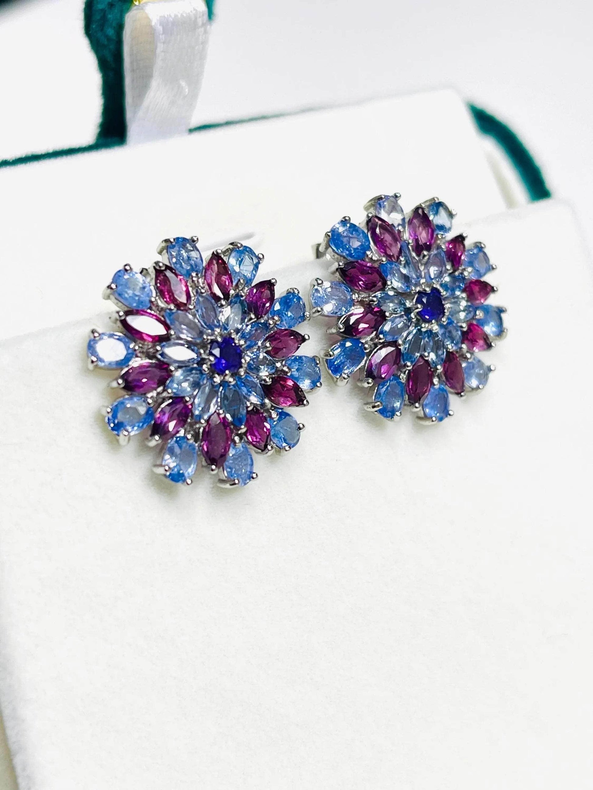Close-up of dazzling tanzanite and rhodolite sunburst earrings on a white display, highlighting gemstone details.