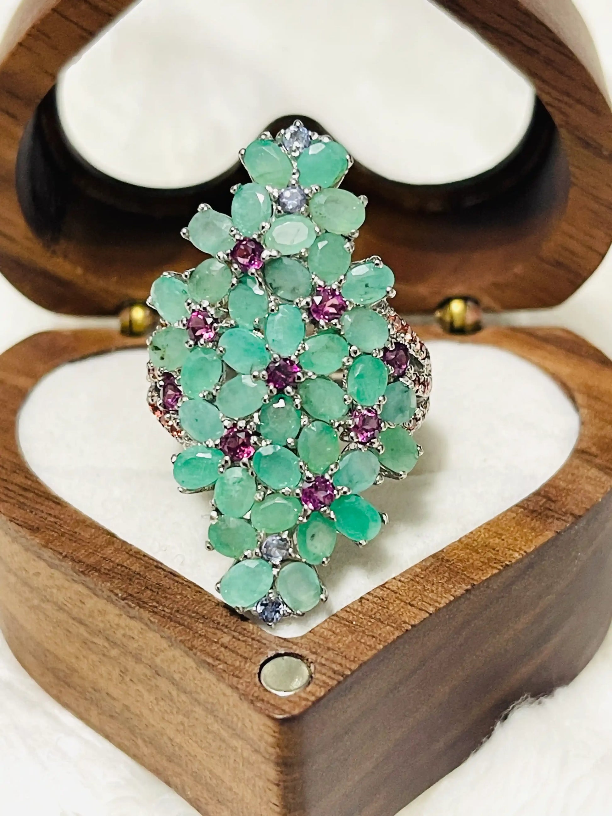 Overhead view of an oversized ring featuring a vibrant cluster of green emeralds and purple amethyst stones in a wooden box.